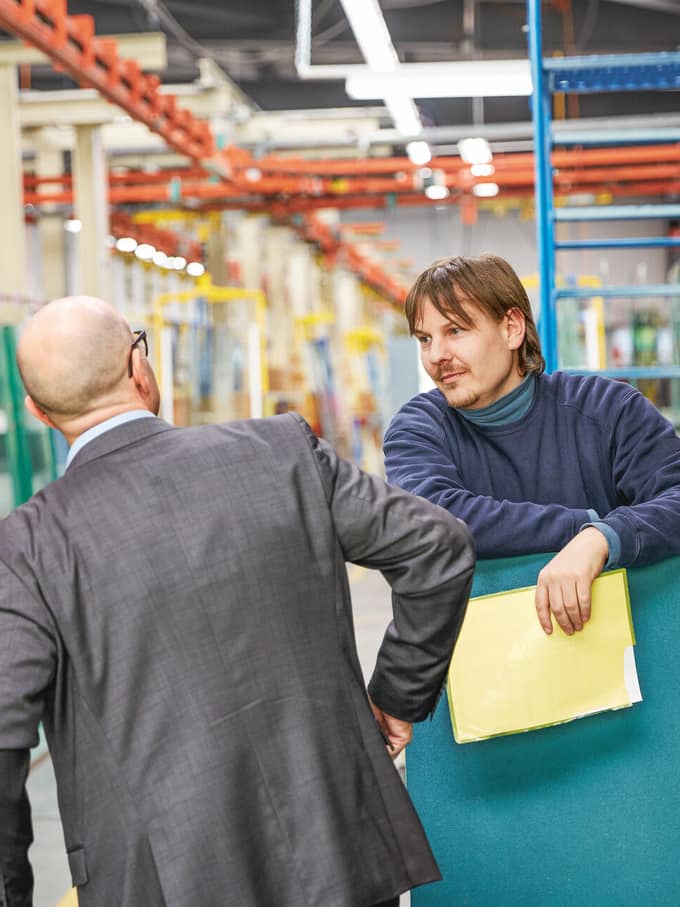 Due uomini in un capannone si guardano. Uno dei due tiene in mano una cartella portadocumenti gialla. Immagine simbolica per la mora nell’accettazione.