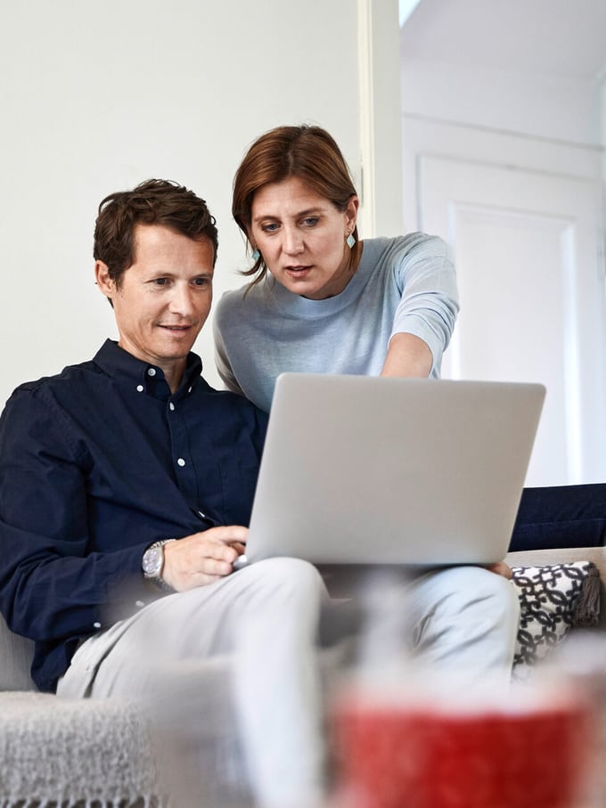 Un uomo è seduto sul divano con il laptop appoggiato sulle gambe. Una donna alle sue spalle indica lo schermo. Rappresentano un’immagine simbolica della continuazione del pagamento del salario e delle prestazioni di indennità giornaliera in caso di incapacità lavorativa.
