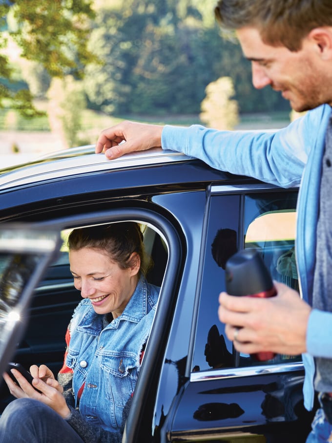 Eine Frau sitzt im Fahrzeug und benutzt ihr Handy, während ein Mann an der Tür steht und sie ansieht. Symbolbild für Handy am Steuer in der Schweiz.