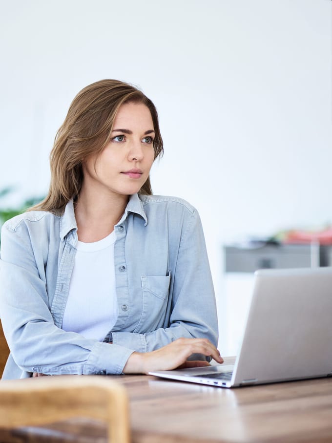 Eine junge Frau sitzt an einem Wohnzimmertisch vor ihrem Laptop mit gedankenversunkenem Blick.