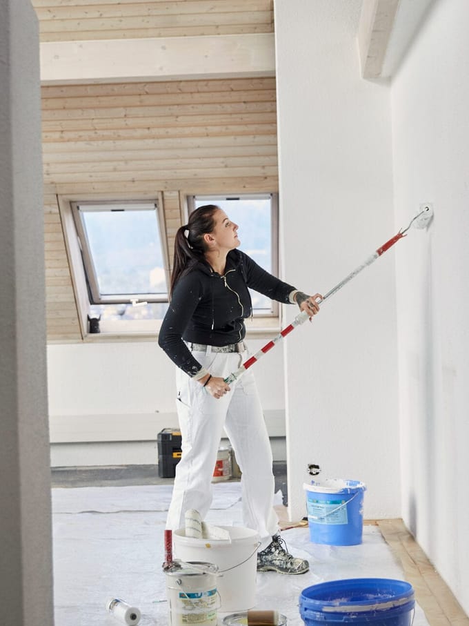 Sur la photo, on voit une peintre de côté. Elle est en train de peindre en blanc un mur avec un rouleau. Autour de vous se trouvent des chaudières de peinture et différents ustensiles.