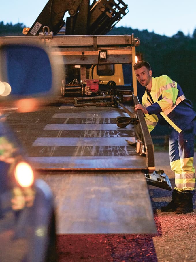 Un collaboratore del servizio di traino carica un veicolo che ha evidentemente avuto una panne. Il proprietario del veicolo osserva la scena da vicino. 