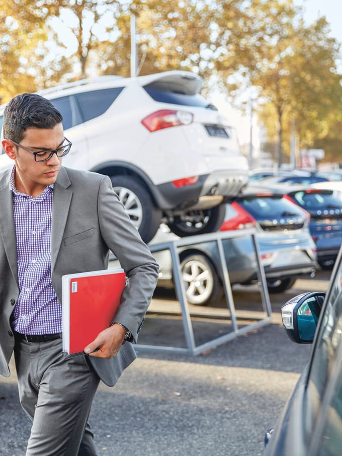 Autoverkäufer läuft über den Parkplatz des Autohauses, er hält eine rote Mappe in der Hand