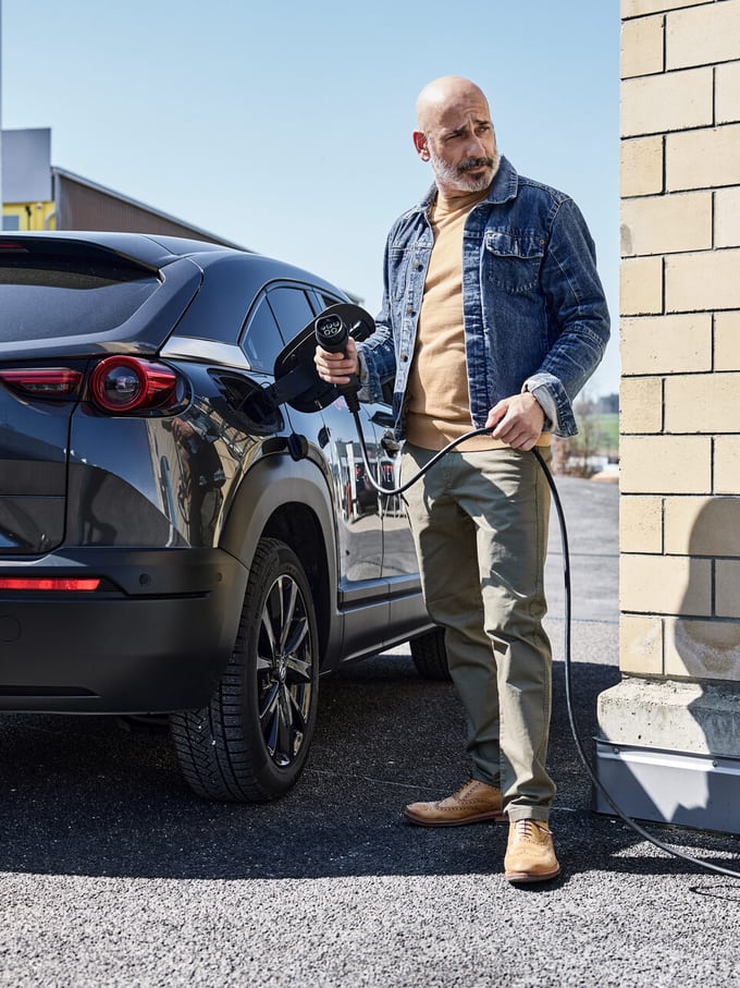 Un homme d’âge moyen, chauve et vêtu d’une veste en jean, se tient prêt à faire le plein avec une station de recharge à côté d’une voiture électrique noire.