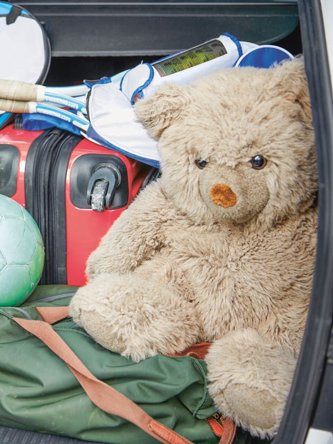 Un ours en peluche est assis dans le coffre d'une voiture, entouré de bagages et de sacs de voyage. Image symbolique de l'annulation d'un hôtel ou d'un voyage et des conditions d'annulation.