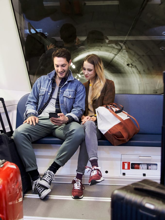 Ein Mann und eine Frau sitzen im hintersten Wagen einer U-Bahn, sie schauen auf das Handy, dass der Mann in der Hand hält. Vor ihnen stehen Personen mit Reisegepäck. Symbolbild für das Thema Datenschutz für kleine Unternehmen.