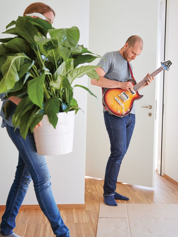 Un homme debout dans le vestibule d’un appartement joue de la guitare pendant qu’une femme portant une plante verte passe à proximité. Image symbolisant la vie commune des couples mariés ou en concubinage.