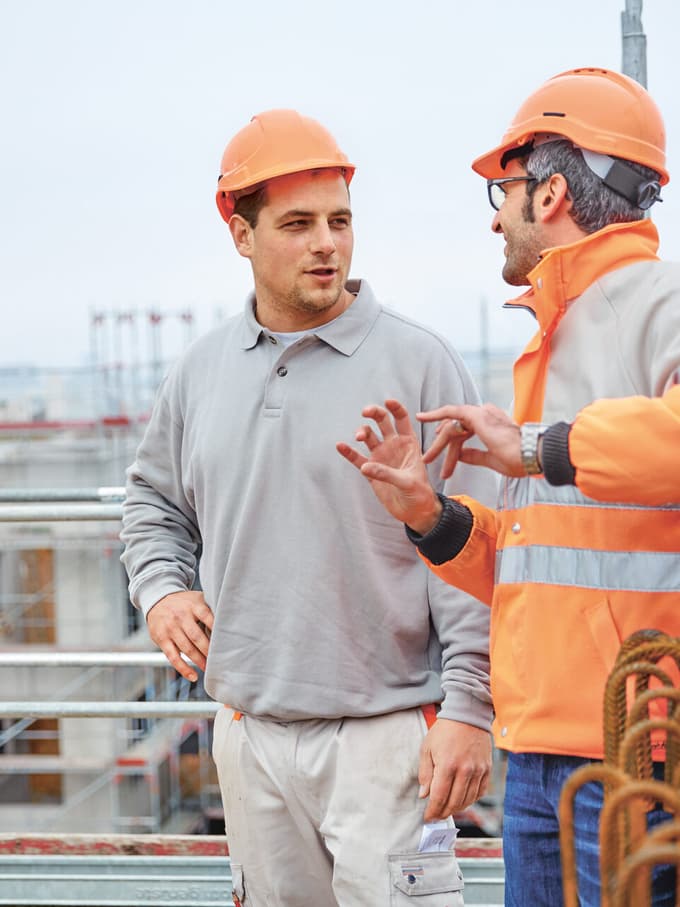 Zwei Männer stehen auf einem Gerüst einer Baustelle mit Bauhüten und besprechen die Bauplanung.