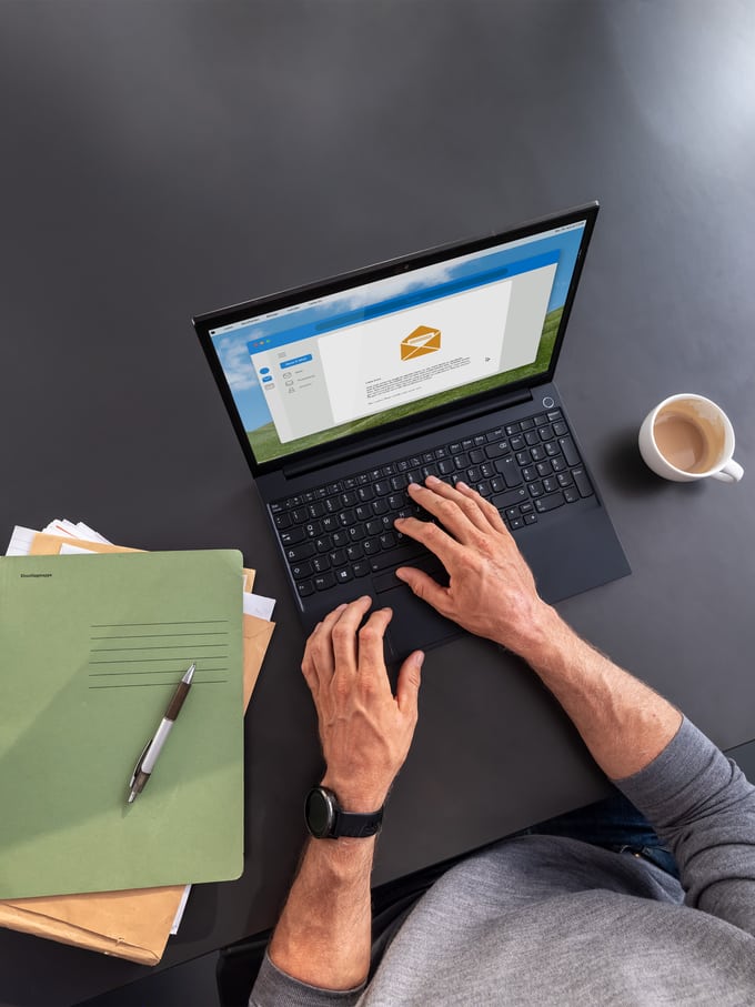 Person bedient einen Laptop auf dem Tisch. Daneben liegen ein Stapel Papier und eine Tasse Kaffee. 