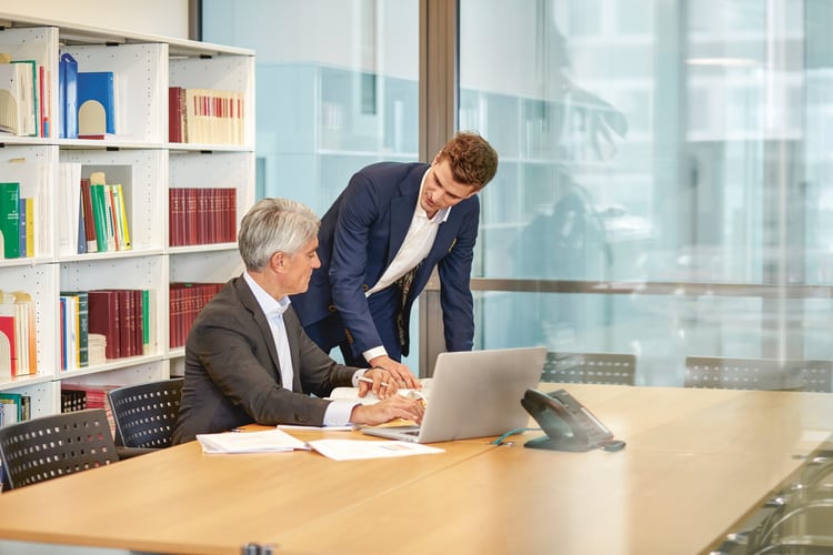 Ein Mann im Anzug sitzt in einem Besprechungszimmer an einem grossen Konferenztisch vor einem Laptop und ausgelegten Dokumenten. Neben ihm steht ein jüngerer Mann im Anzug und bespricht etwas mit ihm. 