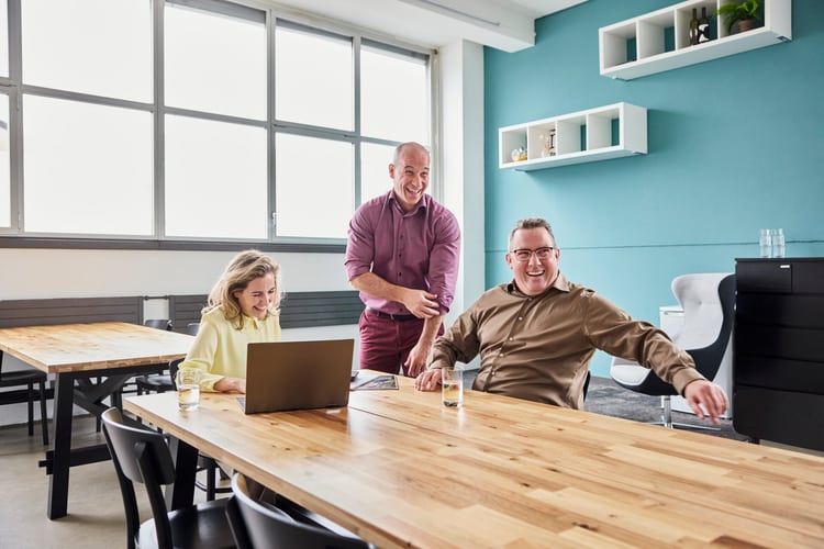 Tre persone sono sedute a un grande tavolo di legno in ufficio e ridono. Una donna e un uomo sono seduti, il terzo uomo è in piedi. Sul tavolo c’è un computer portatile. 