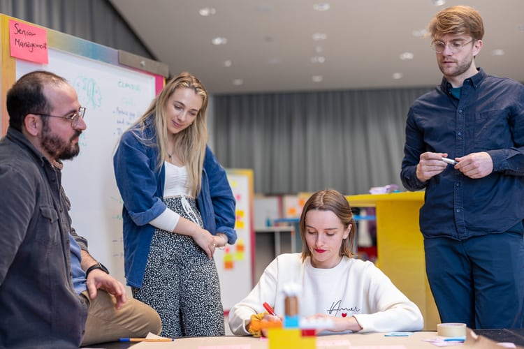Un groupe de participants discute des résultats obtenus. Une femme est assise à la table, les autres se tiennent à sa gauche et à sa droite.
