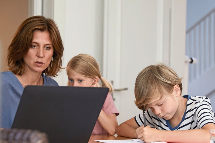 Frau sitzt an Laptop, etwas schein nicht zu stimmen. Kinder daneben eines an den Hausaufgaben, das andere schaut mit auf den Bildschirm.