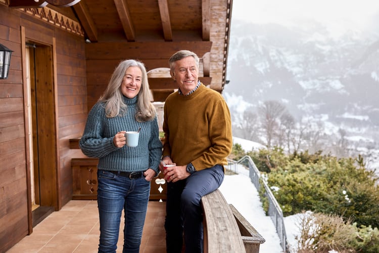 Ein älteres Paar steht auf dem Balkon eines Ferienchalets, lächelt und blickt auf die verschneite Umgebung mit Bergen im Hintergrund. 