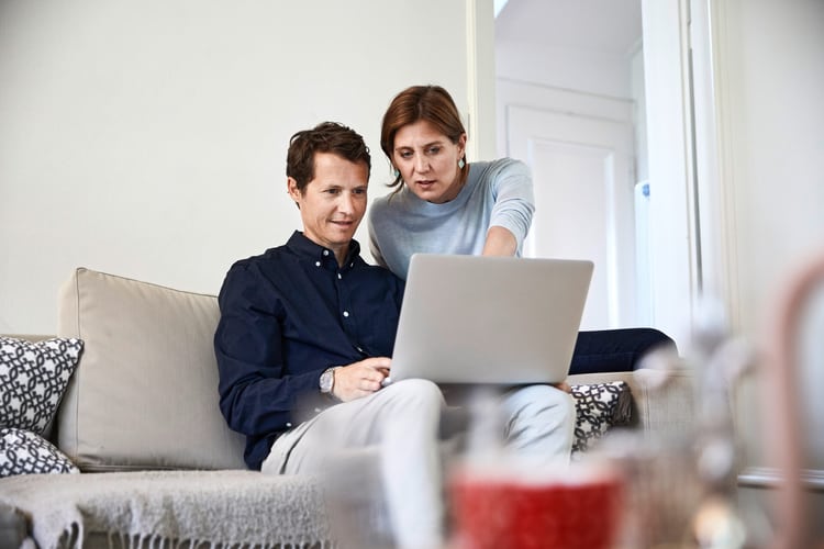 Un uomo è seduto sul divano con il laptop appoggiato sulle gambe. Una donna alle sue spalle indica lo schermo. Rappresentano un’immagine simbolica della continuazione del pagamento del salario e delle prestazioni di indennità giornaliera in caso di incapacità lavorativa.