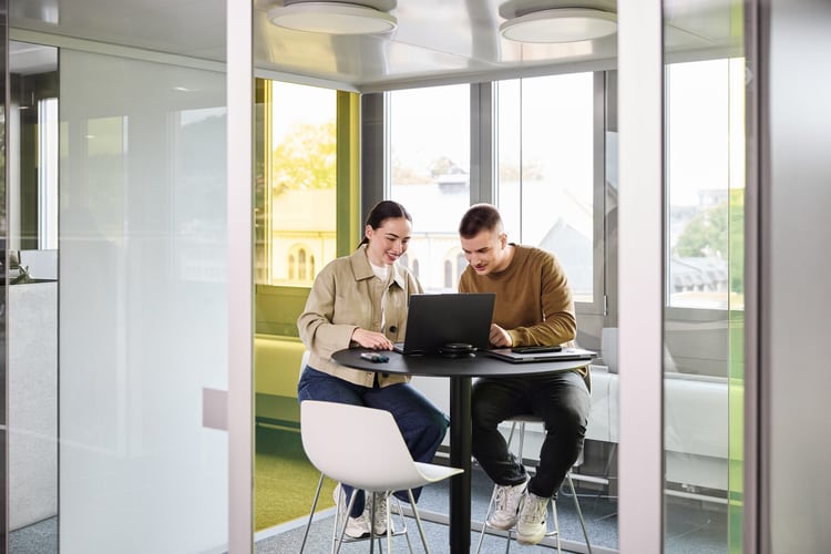 Ein Mann und eine Frau unterhalten sich im Stehen. Eine weitere Frau steht daneben und arbeitet auf ihrem Laptop, der auf einem Stehtisch platziert ist. 