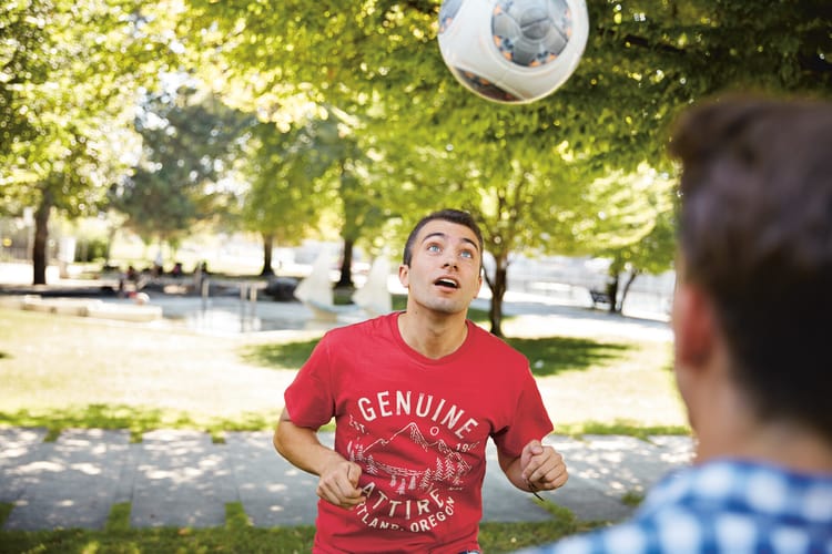 Zwei junge Männer spielen Fussbal auf einer Wiesen mit Bäumen.