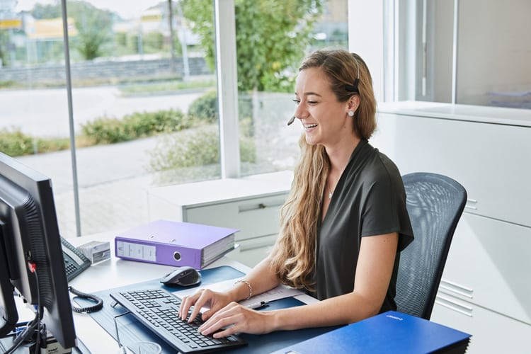 Eine Frau telefoniert mit einem Headset während sie am Computer etwas tippt.