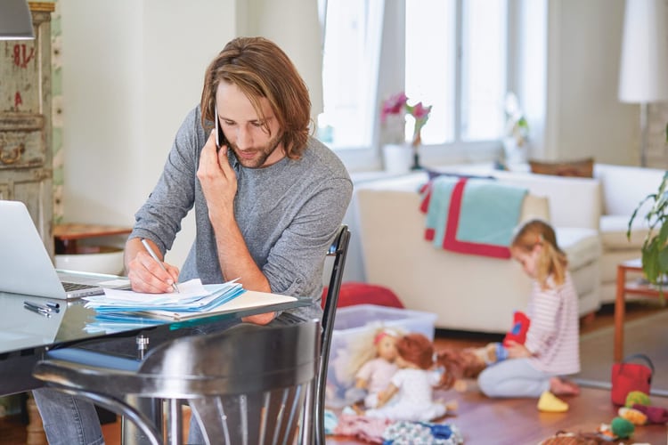 Assis à la table de la cuisine, un homme est en train de téléphoner avec son ordinateur portable et des documents imprimés. Il prend des notes à la main et sa fille joue par terre en arrière-plan.