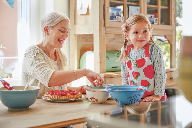 Una madre bionda e il suo bambino siedono insieme a un tavolo della cucina carico di utensili da forno. La madre decora una torta di lamponi raggiante, la bambina indossa un grembiule colorato, si appoggia sul tavolo e sorride.