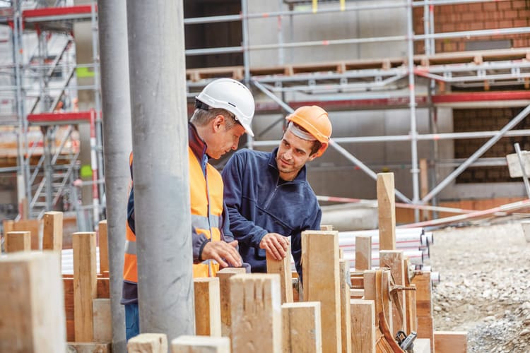 Zwei Männer mit Bauhelmen unterhalten sich auf einer Baustelle vor einer Verschalung für eine Mauer, im Hintergrund erhebt sich ein eingerüstetes Gebäude im Rohbau. 