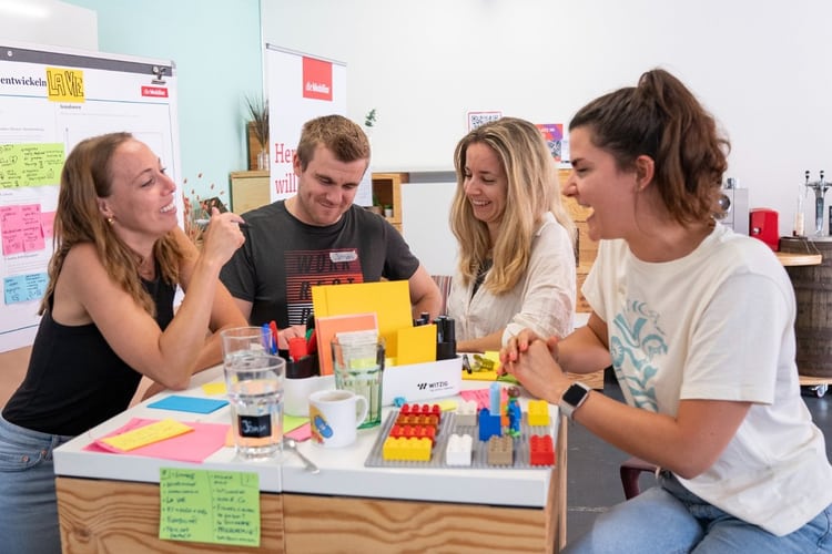 Vier junge Erwachsene sitzen an einem hohen Holztisch, lachen und arbeiten kreativ mit bunten Karten, Stiften und LEGO-Steinen – eine Szene aus einem partizipativen Workshop oder Ideensprint.