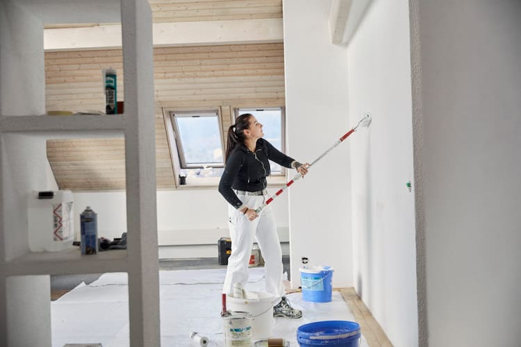 Auf dem Bild sieht man eine Malerin von der Seite. Sie ist dabei eine Wand mit einer Rolle weiss zu streichen. Rund um Sie stehen Farbkessel und verschieden Utensielien.