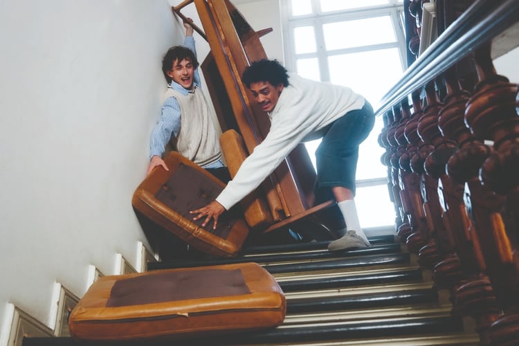 Due giovani uomini manovrano con evidente difficoltà un divano giù per una scala stretta.