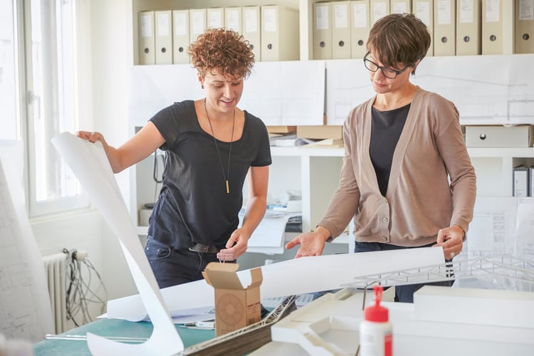 Zwei Frauen stehen an einem Pult und schauen grossformatige Pläne an. Im Hintergrund steht ein Regal, das mit vielen Akten gefüllt ist. 