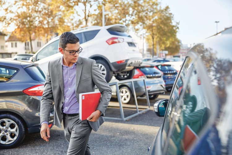 Autoverkäufer läuft über den Parkplatz des Autohauses, er hält eine rote Mappe in der Hand