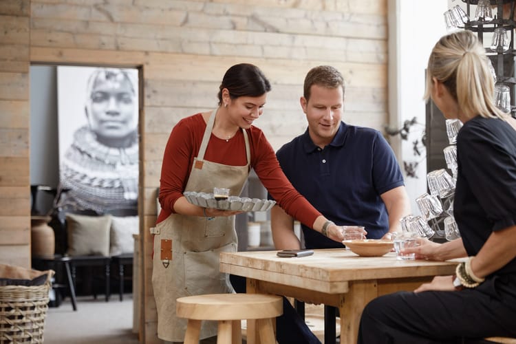 Ein junges Paar sitzt an einem massiven Holztisch in einer Gastwirtschaft im Stil eines Chalets. Eine junge Kellnerin mit rustikaler Schürze stellt ihnen lächelnd Getränke auf den Tisch. 