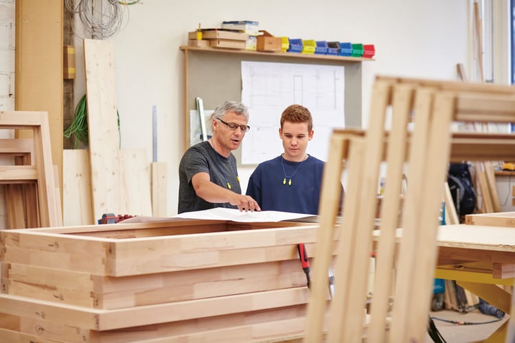 Lehrmeister erklärt in einem Schreinerbetrieb dem Lehrling einen Arbeitsschritt. 