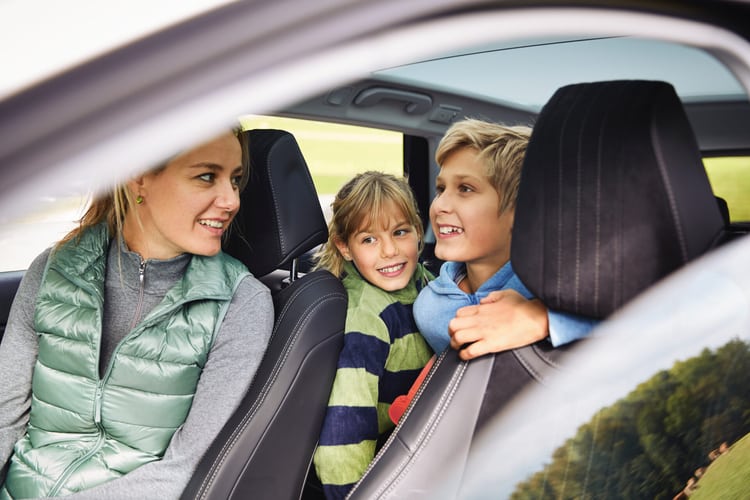 Blick durch das Fahrerfenster in den Innenraum eines Autos: Auf dem Beifahrersitz sitzt eine Mutter, die den Kopf zu ihren fröhlichen Kindern, einem Jungen und einem Mädchen, auf dem Rücksitz wendet und sich lächelnd mit ihnen unterhält. 