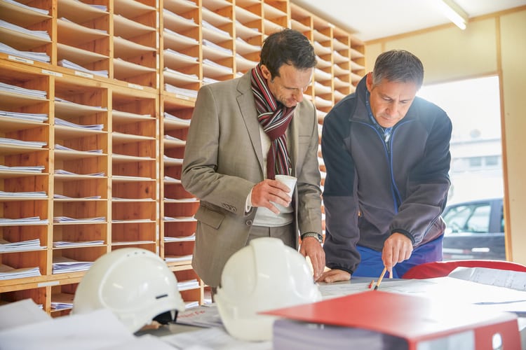 Zwei Männer stehen in einem Baubüro an einem Pult und betrachten Pläne. 