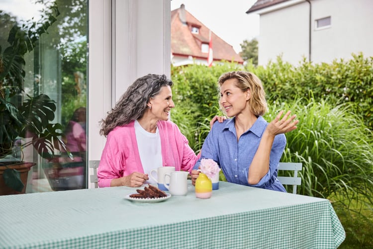 Zwei Frauen unterhalten sich auf einer Terrasse an einem Gartentisch.