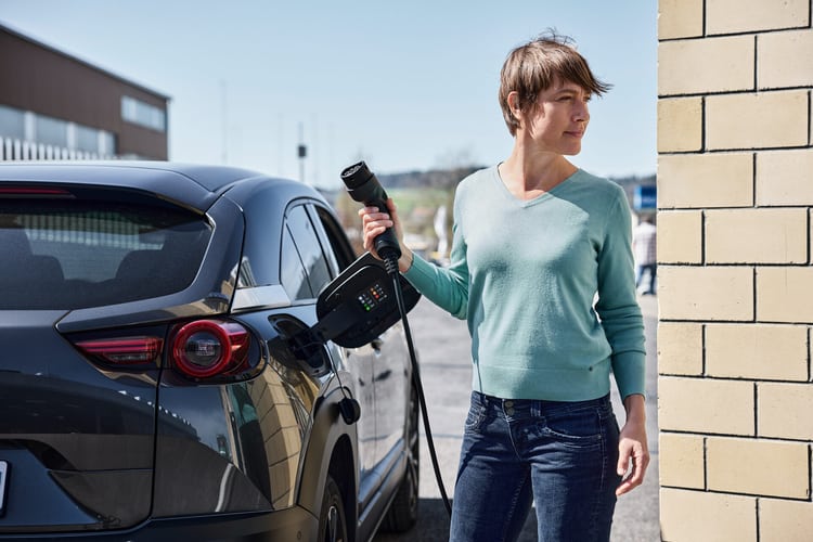 Une femme se tient debout à côté de sa voiture électrique, dont le clapet de recharge est ouvert. Elle tient à la main un câble de recharge. 