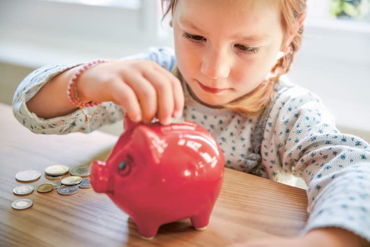 Ein kleines Mädchen sitzt an einem Tisch mit einigen Münzen und steckt eine von ihnen in ein Sparschwein. 