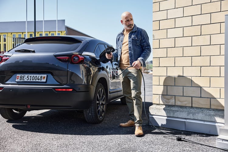 Un homme d’âge moyen, chauve et vêtu d’une veste en jean, se tient prêt à faire le plein avec une station de recharge à côté d’une voiture électrique noire.