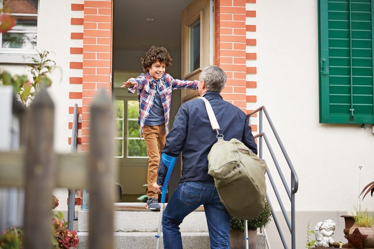 Un homme d’âge moyen se dirige vers une entrée avec des béquilles. Un garçon en chemise à carreaux l’accueille à bras ouverts.