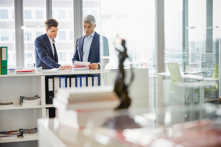 Un homme examine des documents posés sur un meuble de bureau. Le premier plan est en partie occupé par une statuette aux contours flous. L’image symbolise le guide sur le contrat d’entreprise.