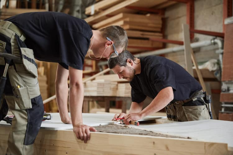 Zwei Schreiner arbeiten in einer Zimmerei an einem Bauteil aus Holz, im Hintergrund sind Bretter entlang der Wand gestapelt. 