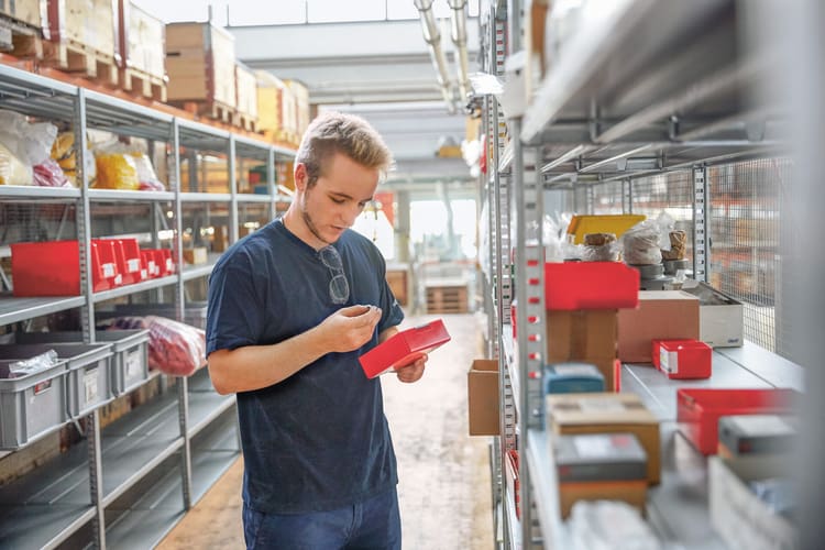 Ein junger Mann steht vor einem Hochregal und hält eine rote Kiste in den Händen. Er holt daraus einen Gegenstand heraus. 