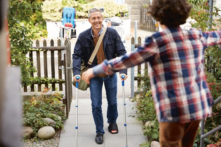 Ein Mann mit Krücken wird zu Hause vom Sohn begrüsst.