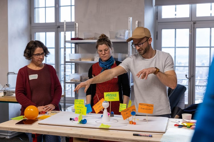 Un gruppo di partecipanti discute i risultati ottenuti. Due donne e un uomo sono in piedi attorno a un tavolo, su cui presentano il modello elaborato. 