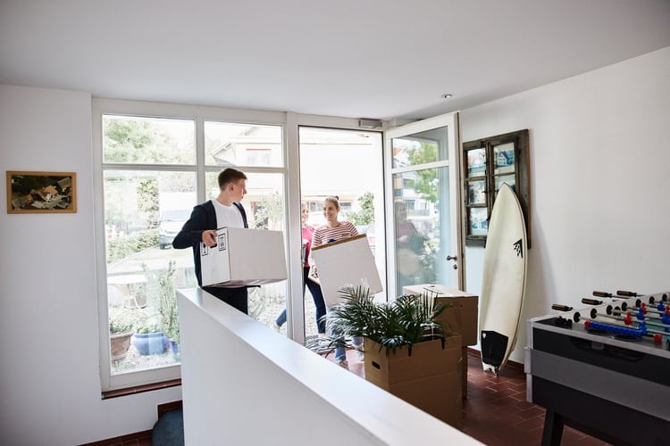 Zwei Personen betreten ein Haus mit einer Kartonkiste. Im Vordergrund stehen ein Surfbrett sowie eine Kiste mit einer Pflanze. Symbolbild für Mietkaution.