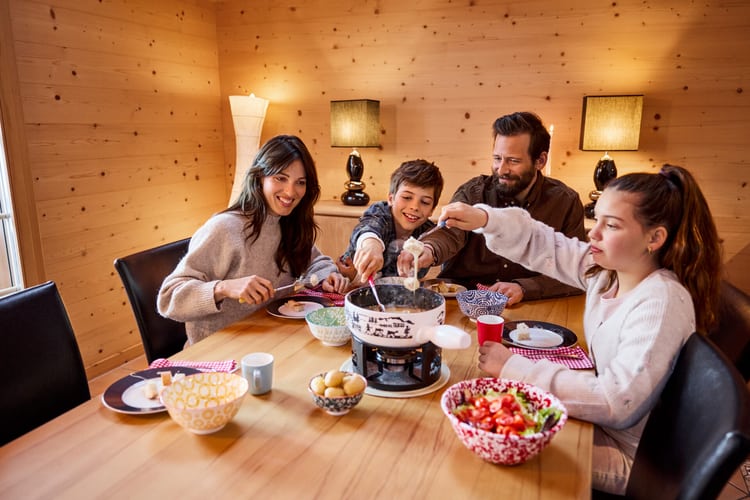 Eine Familie mit Mutter, Vater, Junge und Mädchen sitzt am Tisch eines Ferienchalets mit Holzwänden und geniesst ein Käsefondue. 