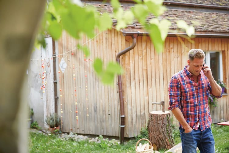 Mann im karierten Flanellhemd steht vor einem Holzhaus und spricht in ein Smartphone. Im Hintergrund sieht man einen Baumstumpf mit einer Axt. Symbolbild für Kapprecht.