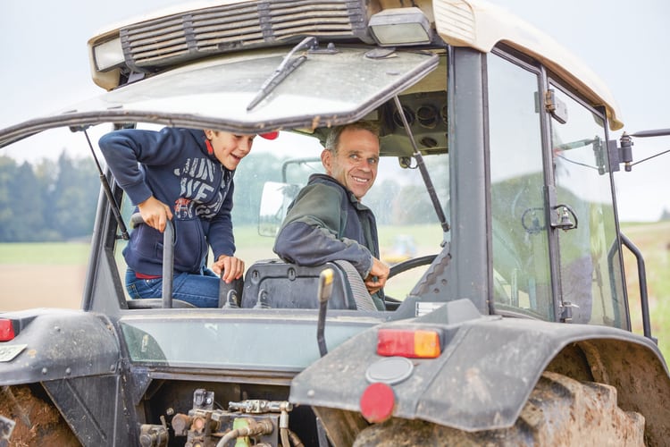 Blick von hinten in die geöffnete Führerkabine eines Traktors, wo ein Landwirt und sein Sohn sitzen und fröhlich in die Kamera blicken. 