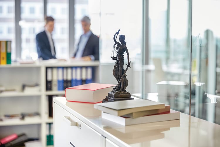 In einem Grossraumbüro steht im scharfen Vordergrund auf einem Korpus eine kleine Justitia-Statue mit Waage und Schwert. Im unscharfen Hintergrund diskutieren zwei Männer miteinander. 