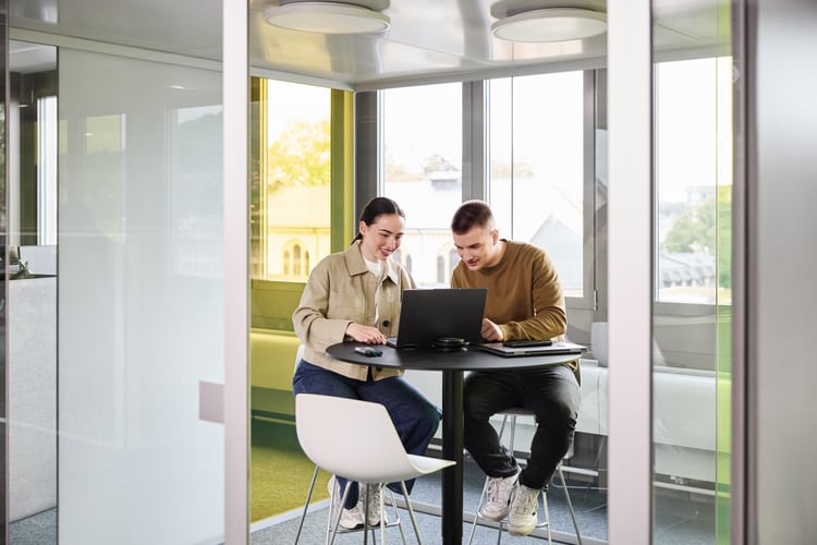 Ein Mann und eine Frau unterhalten sich im Stehen. Eine weitere Frau steht daneben und arbeitet auf ihrem Laptop, der auf einem Stehtisch platziert ist. 